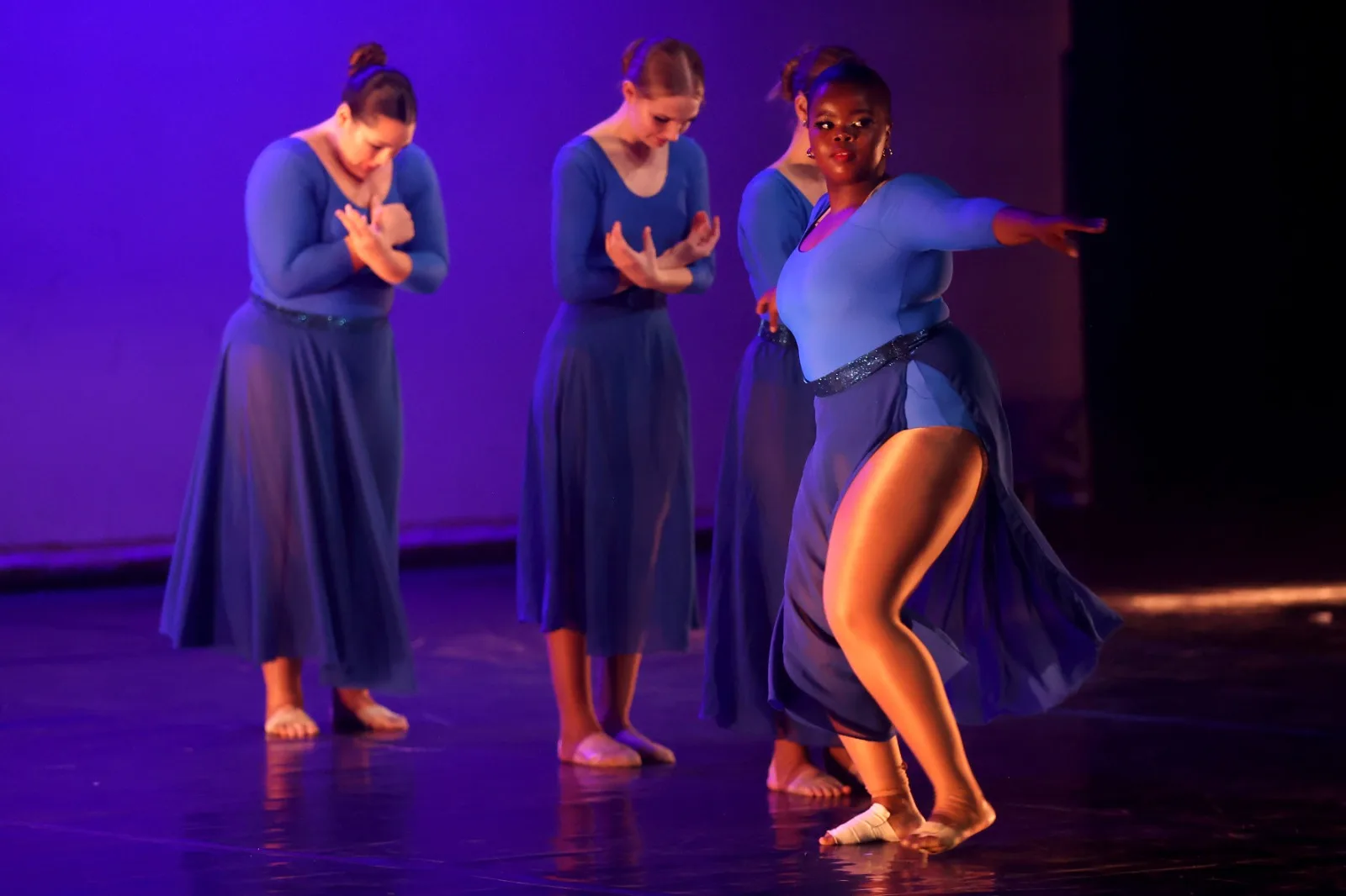 Three JJDC dancers in blue contemporary costumes with hands clasped, a fourth reaching outward, under deep blue stage light