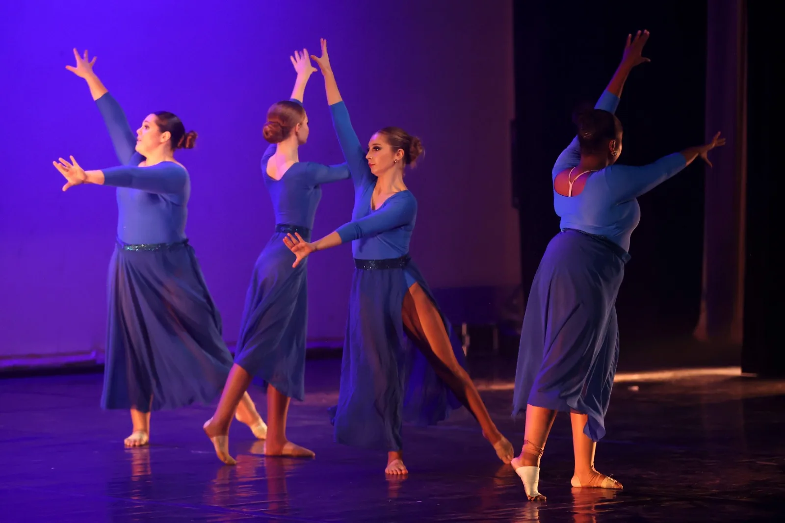 Four JJDC dancers in blue contemporary costumes reaching upward together under purple stage light