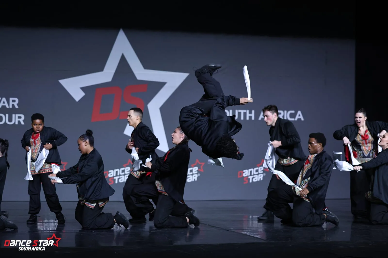 A JJDC hip hop dancer captured mid-backflip on stage at the Dance Star South Africa tour, teammates kneeling in formation behind