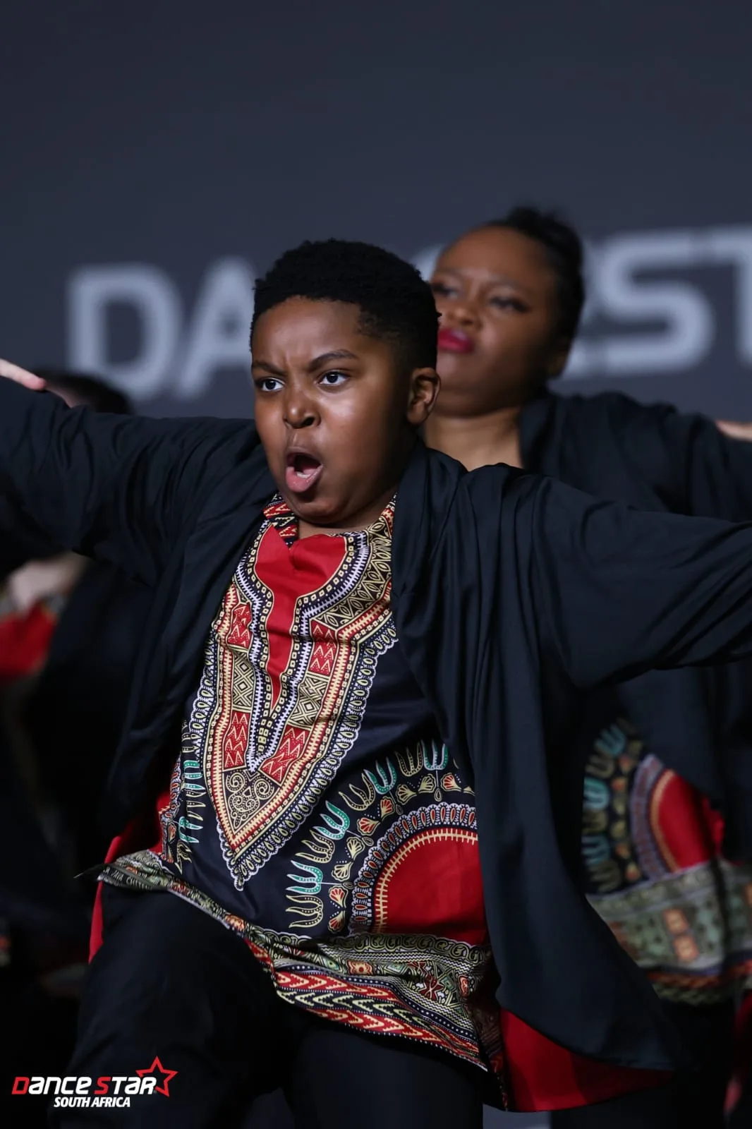 A young dancer in a Dashiki-print jacket, mouth open mid-shout, performing a hip hop routine
