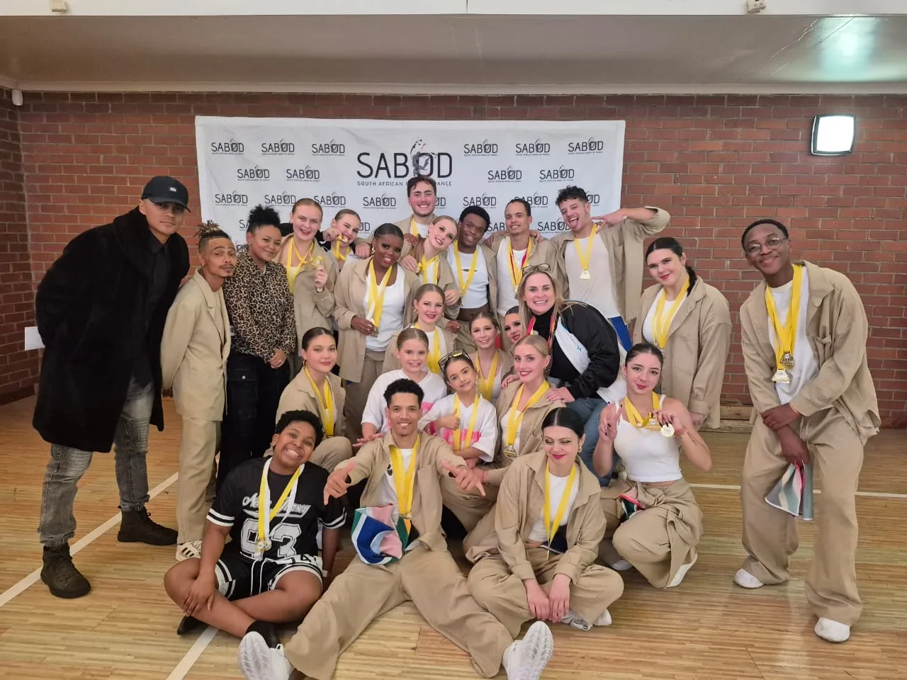 JJDC dancers in beige tracksuits with medals posing backstage at a SABOD competition