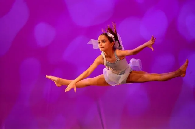 A young dancer captured mid-leap on stage under coloured lighting